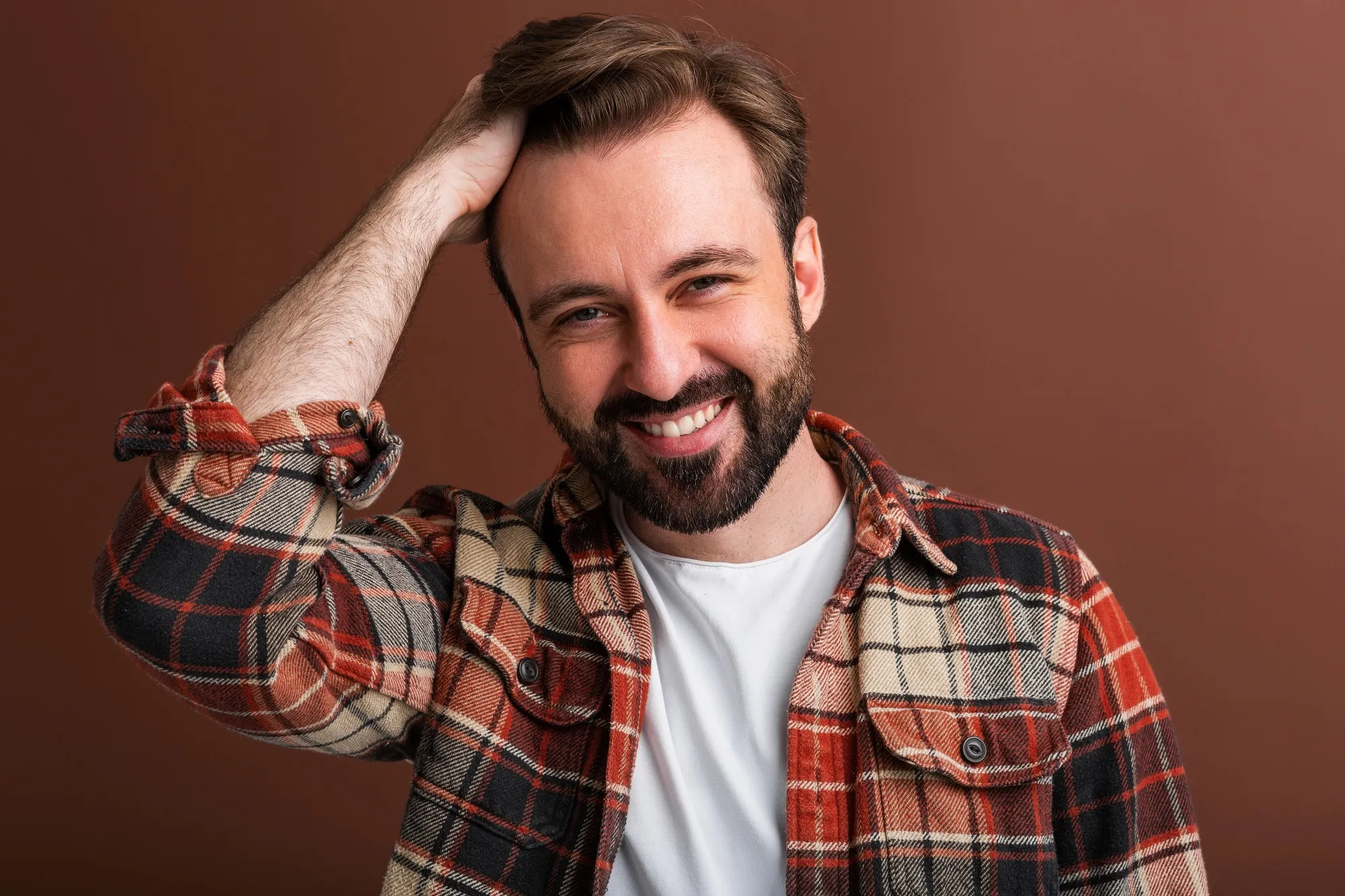Lachende man met een baard en kort, bruin haar strijkt met zijn hand door zijn haar, gekleed in een geruit overhemd over een wit T-shirt.